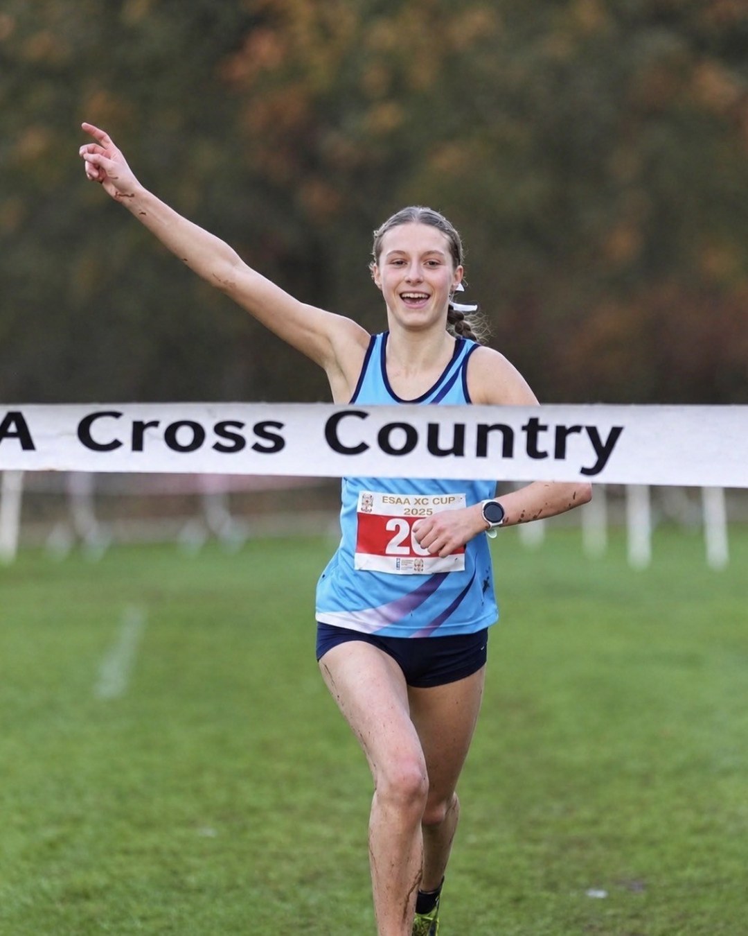 Kitty Scott, in a light blue vest and navy shorts, breaking the tape with one arm raised to win an English Schools' Athletic Association cross country race, her legs splashed with mud