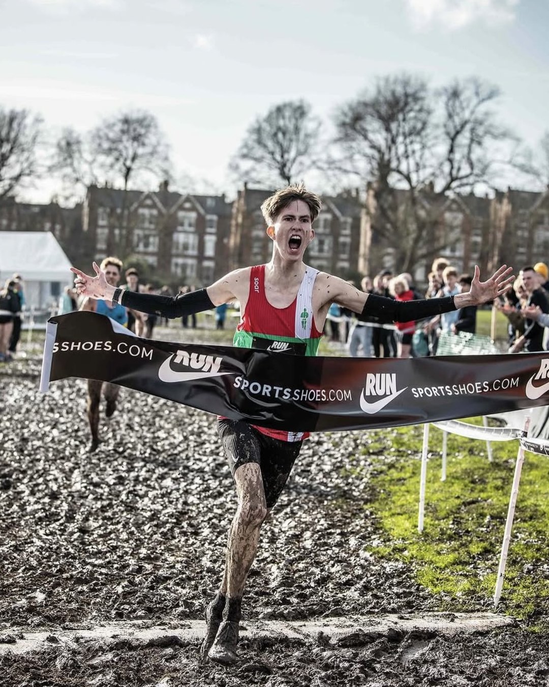 Matt Pickering crossing the line first at the English National Cross Country Championships, arms spread wide and his legs covered in mud, breaking a sportsshoes.com finish tape